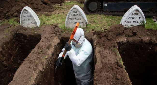 Работник в защитном костюме роет могилы предполагаемым жертвам COVID-19